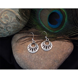 May include: A pair of silver-colored, openwork earrings with a floral design. The earrings are circular with a series of petal-shaped cutouts around the edge. They hang from simple ear wires. The earrings are displayed on a brown stone with a peacock feather in the background.