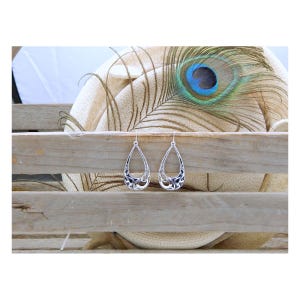 May include: A pair of silver teardrop earrings with an open filigree design. The earrings are displayed on a rustic wooden surface, with a peacock feather and a neutral-toned background.