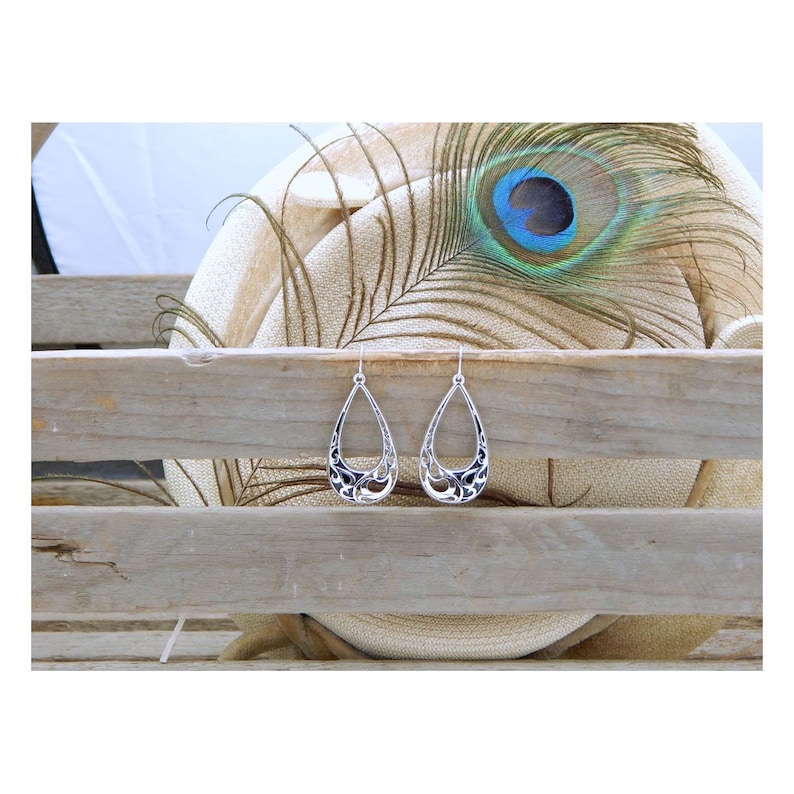 May include: A pair of silver teardrop earrings with an open filigree design. The earrings are displayed on a rustic wooden surface, with a peacock feather and a neutral-toned background.