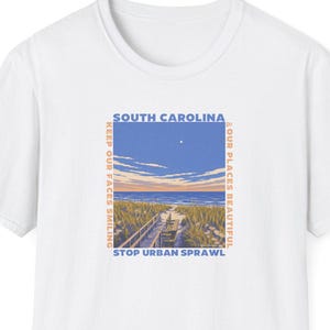 Op de afbeelding: Een wit t-shirt met een rechthoekige afbeelding van een strand in South Carolina met een houten vlonderpad, zandduinen en een blauwe lucht. De tekst luidt SOUTH CAROLINA, KEEP OUR FACES SMILING, OUR PLACES BEAUTIFUL en STOP URBAN SPRAWL.