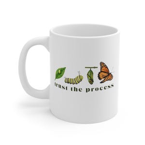 May include: White ceramic mug featuring a watercolor illustration of a butterfly life cycle. The design includes a leaf, caterpillar, chrysalis, and butterfly, with the words "trust the process" in black text.