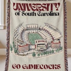 May include: A woven throw blanket featuring a beige background with a detailed illustration of the University of South Carolina stadium. The text reads UNIVERSITY of South Carolina at the top and GO GAMECOCKS at the bottom with fringed edges.