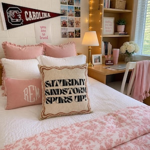 May include: A square throw pillow with a beige fabric cover featuring a scalloped brown border and the black text SATURDAY SANDSTORM SPURS UP! centered on the front. The pillow is placed on a white bedspread with pink pillows and a pink floral throw.
