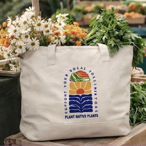 Puede incluir: Un bolso tote de color crema con el texto "Apoya a tus polinizadores locales, planta plantas nativas". La bolsa está llena de margaritas blancas, flores naranjas y hierbas verdes. La bolsa está sobre una superficie de madera.