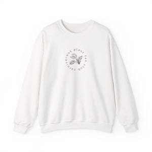 May include: A white long-sleeve crewneck sweatshirt featuring a black circular graphic on the chest. The design includes a detailed illustration of a Venus flytrap plant surrounded by the text "carnivorous plant fan club" in a circular font.