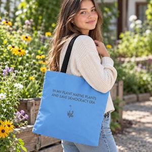 May include: A light blue tote bag with black straps featuring the text IN MY PLANT NATIVE PLANTS AND SAVE DEMOCRACY ERA printed in black, accompanied by a small, simple black line drawing of a single plant centered below the text.