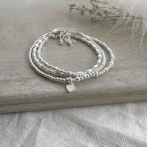May include: A set of three silver-toned beaded bracelets featuring a small heart-shaped charm engraved with the letter M. The bracelets are constructed from small, uniform metallic spheres and elongated beads, connected by a silver-toned clasp mechanism.
