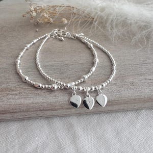 May include: Two silver beaded bracelets, each with heart-shaped charms bearing engraved initials. The bracelets are presented on a light wooden surface. The background is soft and out of focus.