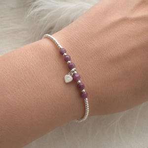 May include: A bracelet with small, round, purple beads and silver beads. A silver heart charm with a letter dangles from the bracelet. The bracelet is worn on a wrist. The beads are approximately 0.5 cm in diameter.