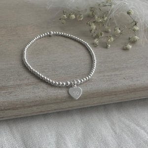 May include: A silver beaded stretch bracelet featuring a small heart-shaped charm. The charm is engraved with the number 30. The bracelet is composed of numerous small, uniform, polished silver-coloured spherical beads arranged in a circular loop.