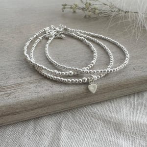 May include: A set of three silver-toned beaded bracelets featuring small, uniform metallic spheres. One bracelet includes a small, dangling heart-shaped charm engraved with the letter M. The bracelets are arranged in a circular, layered formation.