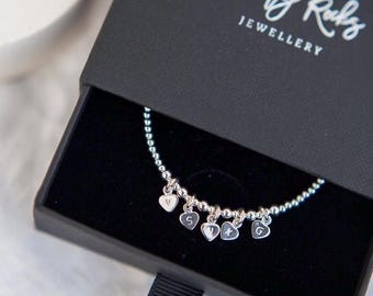May include: A silver beaded bracelet with five small heart-shaped charms, each engraved with a letter, resting inside a black sliding jewelry box. The box lid features the white printed text Dainty Rocks JEWELLERY with a feather graphic above it.