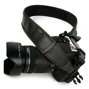 May include: A black leather camera strap with red stitching and the word "ACTION" in gold lettering. The strap is attached to a black DSLR camera with a lens.