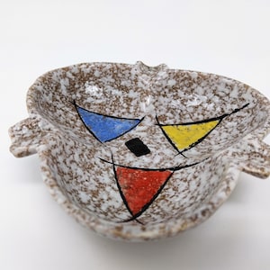 Könnte beinhalten: Ein weißer Aschenbecher aus Keramik mit brauner, gesprenkelter Glasur. Der Aschenbecher hat ein geometrisches Design, das in Blau, Gelb, Rot und Schwarz bemalt ist.