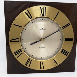 May include: A gold-toned clock with Roman numeral markings on a brown wooden base. The clock face reads "JUNGHANS electronic ATO-MAT".