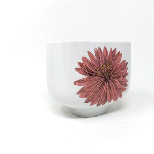 Puede incluir: Cuenco de cerámica blanca con forma cuadrada redondeada. Una gran flor rosa detallada con estambres amarillos está impresa en el lateral. El cuenco tiene una pequeña base circular.