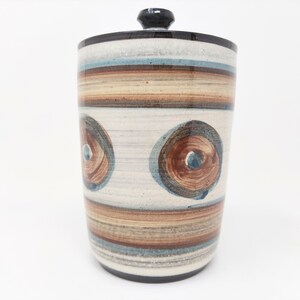 May include: A white ceramic jar with a black lid and a brown, blue, and white striped pattern. The jar has two large brown circles with blue centers.
