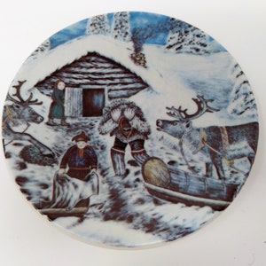May include: A white ceramic plate with a winter scene featuring a small wooden cabin, two people, and two reindeer pulling a sled. The scene is painted in brown, blue, and white.