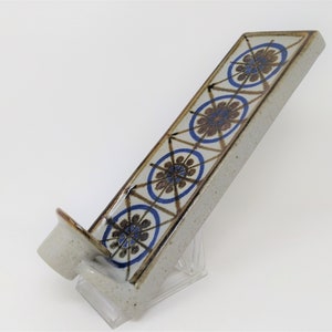 Puede incluir: Un portavelas de pared de cerámica con un patrón geométrico azul y marrón. El portavelas tiene una sola ranura para velas y está diseñado para montarse en una pared.