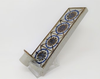 Puede incluir: Un portavelas de pared de cerámica con un patrón geométrico azul y marrón. El portavelas tiene una sola ranura para velas y está diseñado para montarse en una pared.