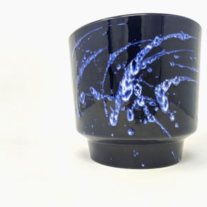 May include: A dark navy blue ceramic cup featuring a glossy finish and a decorative pattern of bright blue paint splatters that streak across the surface. The cup has a wide, slightly flared rim and a narrower, cylindrical base for stability.