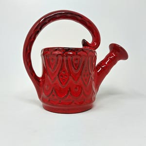Könnte beinhalten: Eine rote Keramik-Gießkanne mit einem dekorativen Design. Der Griff ist in Form eines Vogelkopfes gestaltet.