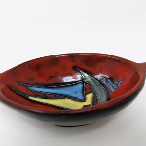 May include: A red ceramic dish with a black rim and a colorful abstract design. The dish has a handle on one side.
