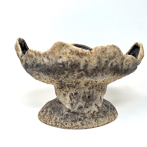 Op de afbeelding: Een getextureerde keramische kom met een voet. De kom heeft een brede, golvende rand met donkere accenten en een neutraal kleurenpalet van beige en bruin. De binnenkant is donker. De kom is waarschijnlijk handgemaakt.