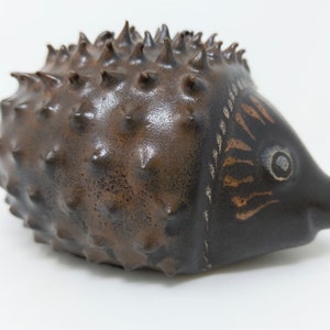 May include: A brown ceramic hedgehog figurine with a textured, spiky back and a painted face with a black eye.