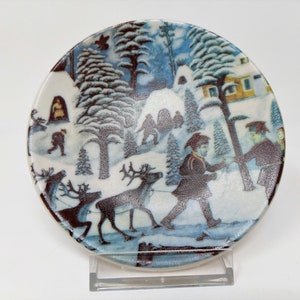 May include: A small ceramic plate with a winter scene painted on it. The scene features a snowy forest with houses, trees, and people walking with reindeer. The plate is white with blue and brown accents.