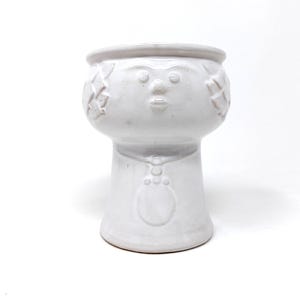 May include: White ceramic planter with a face design. The planter has a round top, a face with a nose and lips, and decorative elements on the sides. The base is a pedestal shape.