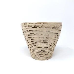 May include: A beige ceramic planter with a textured, woven-like pattern. The planter has a wide opening and a slightly tapered shape. The design features horizontal and vertical lines in a darker shade, creating a basket-weave effect. The planter is suitable for indoor or outdoor use.