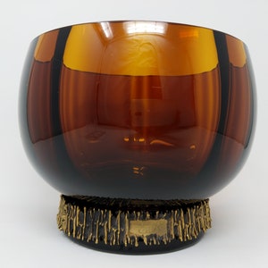 Puede incluir: Un cuenco de vidrio marrón con una base de metal dorado. La base tiene un diseño texturizado y una pequeña etiqueta con la letra "B".