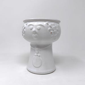 May include: A white ceramic planter with a face design. The planter has a round bowl shape with a face sculpted into the side. The base is a pedestal shape with a rounded bottom. The planter is a solid white color.