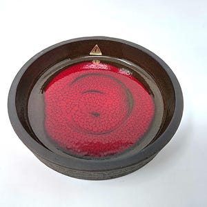 May include: A round, dark brown ceramic bowl with a vibrant red interior. The red interior features a textured, swirling pattern. A small, gold triangle is visible on the rim. The bowl is likely for decorative or serving purposes.