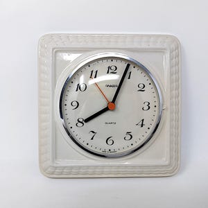 May include: A square cream-coloured wall clock with a textured border and a circular face. The clock features black Arabic numerals, black hour and minute hands, a thin orange second hand, and the text Ankra and Quartz printed on the face.