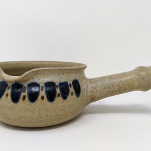 May include: A beige ceramic gravy boat with a brown and blue patterned design. The boat has a handle and a spout.