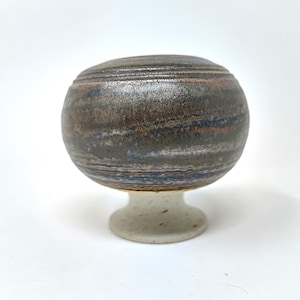 Könnte beinhalten: Eine handgefertigte Keramik-Sockelschale. Die Schale hat eine abgerundete Oberseite mit horizontalen Streifen in Braun-, Blau- und Grautönen. Sie ruht auf einem kurzen, gesprenkelten, cremefarbenen Sockel. Die Schale steht vor einem weißen Hintergrund.