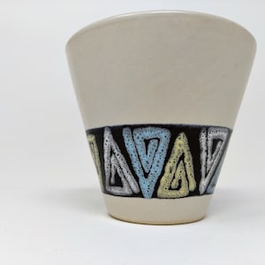 Op de afbeelding: Een witte keramische plantenpot met een zwart, blauw en geel geometrisch patroon rond de rand.