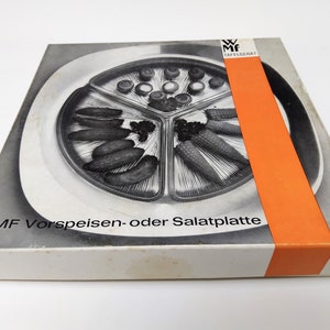 May include: A white box with an orange stripe on the side. The box contains a silver serving platter with three sections. The platter is filled with various foods, including grapes, olives, and vegetables. The box has the text "WMF Vorspeisen- oder Salatplatte" printed on the front.