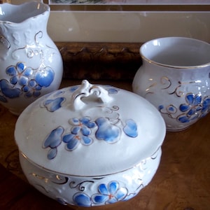 May include: A white porcelain bathroom set with a blue and gold floral design. The set includes a pitcher, a mug, and a covered bowl.