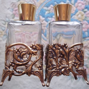 May include: Two clear glass perfume bottles with gold tops and ornate gold metal stands. The stands have a floral design.