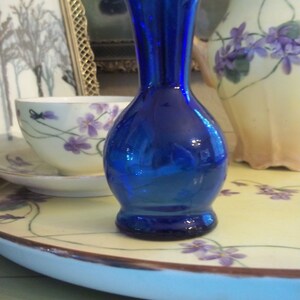May include: A cobalt blue glass vase with a round base and a narrow neck. The vase is sitting on a white plate with a floral design.