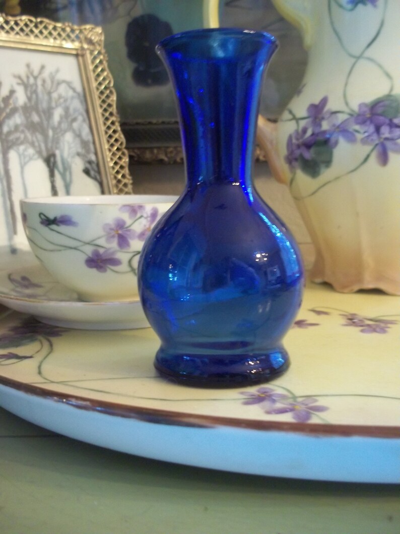 May include: A cobalt blue glass vase with a round base and a narrow neck. The vase is sitting on a white plate with a floral design.