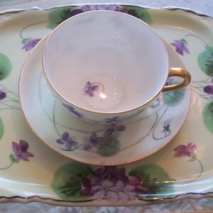 May include: A white porcelain teacup and saucer set with a delicate floral design of purple violets and green leaves. The set is on a matching tray with a scalloped edge.