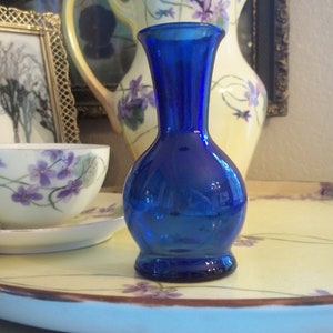 May include: A cobalt blue glass bud vase with a rounded body and a flared top. The vase is sitting on a white tray with a floral design.