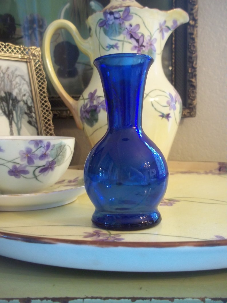May include: A cobalt blue glass bud vase with a rounded body and a flared top. The vase is sitting on a white tray with a floral design.