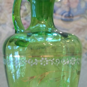 May include: A green glass bottle with a white and gold design. The bottle has a handle and a round top.
