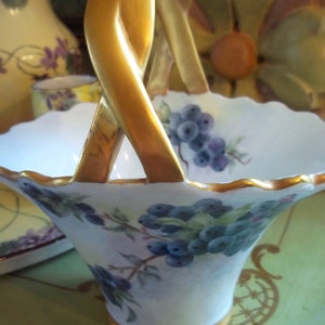May include: A white porcelain basket with a gold handle and a painted design of blue berries and green leaves. The basket is sitting on a table with a green surface.