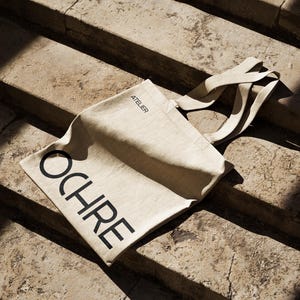 May include: A beige canvas tote bag with the word "OCHRE" printed in large, black capital letters. The word "ATELIER" is printed in smaller letters above the word "OCHRE". The bag has long, beige straps and is resting on stone steps.